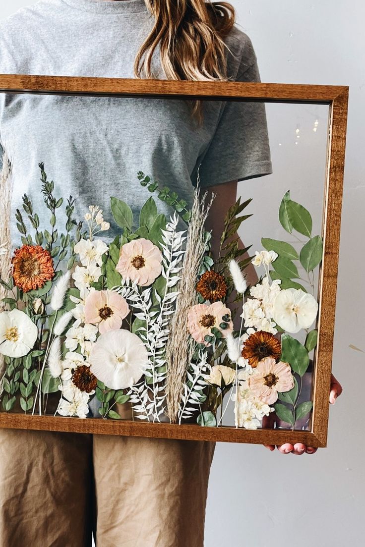 a woman holding up a framed painting with flowers and leaves on it's sides