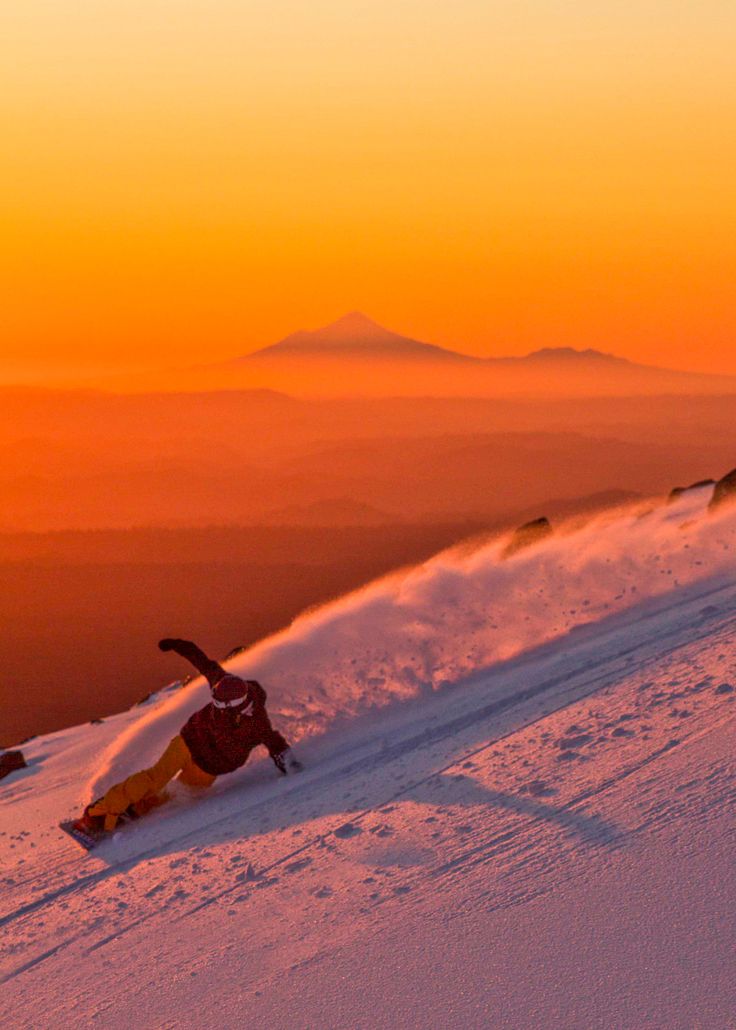 a man riding a snowboard down the side of a snow covered slope at sunset