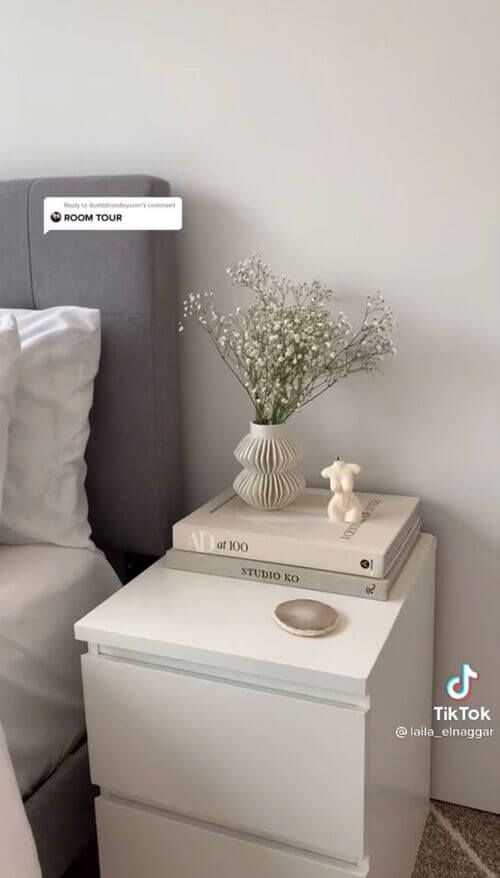 a nightstand with two books on it next to a bed and flowers in a vase