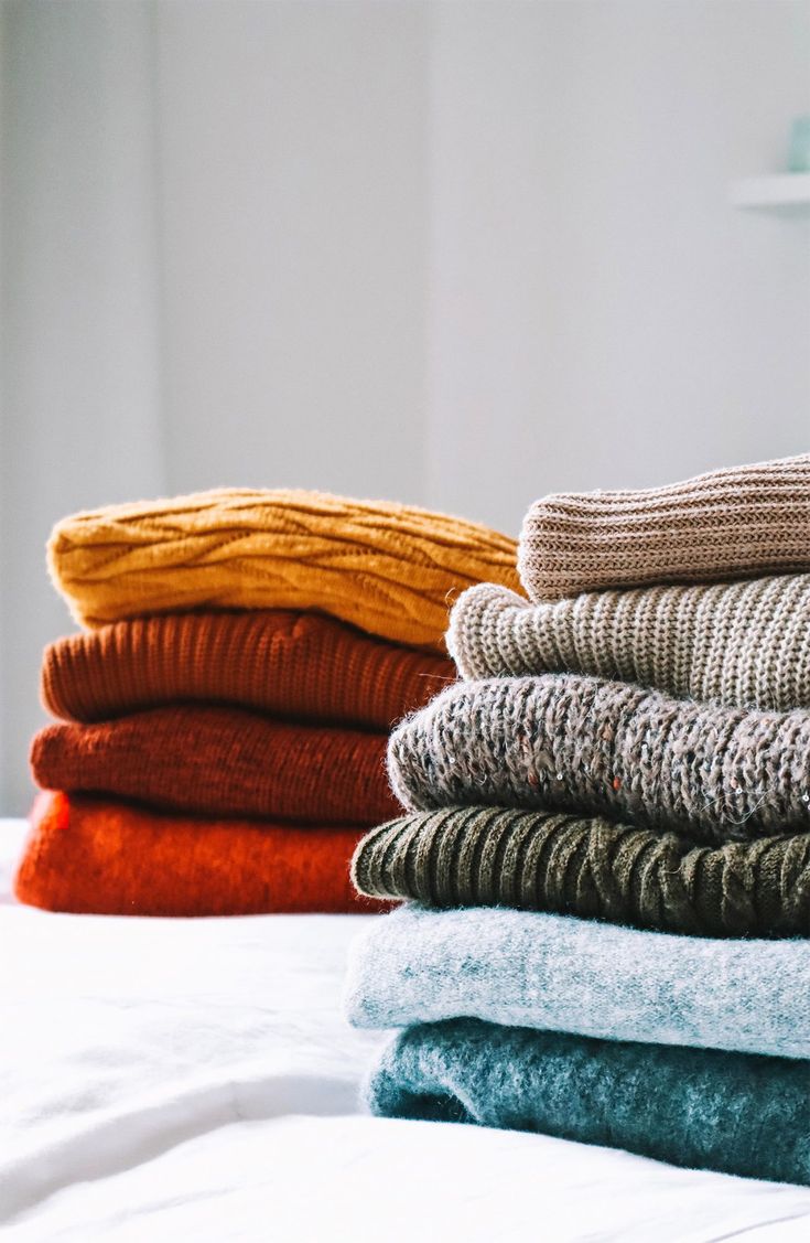 stack of folded sweaters on top of a bed with white sheets and pillows in the background