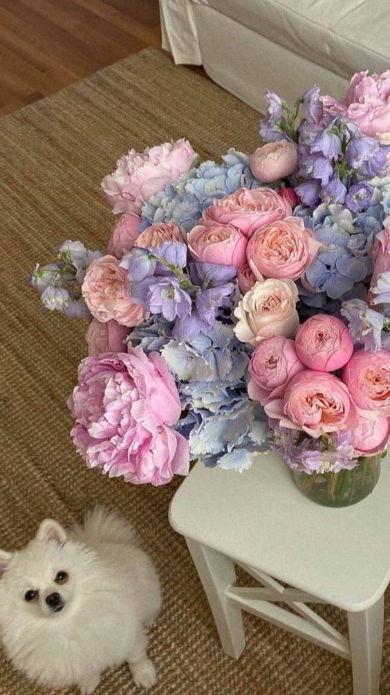 a small white dog sitting on the floor next to a vase filled with pink and blue flowers