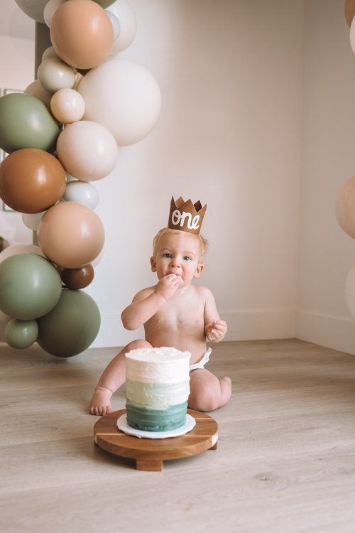 a baby sitting on the floor with a cake in front of him and balloons behind him