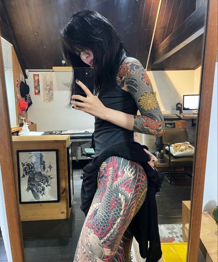 a woman with black hair and tattoos is looking at her cell phone in the mirror