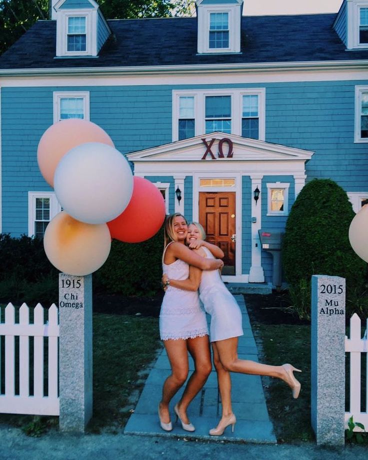 two women hugging each other in front of a blue house with white picket fence and balloons