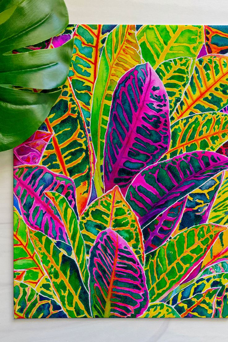 a painting of colorful leaves on a white surface next to a green leafy plant