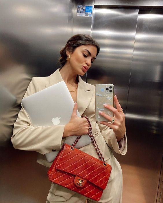 a woman holding an apple laptop computer in front of a silver wall with a red purse on it