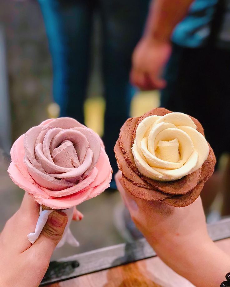 two people holding up small pastries with frosting on them, one is pink and the other is yellow