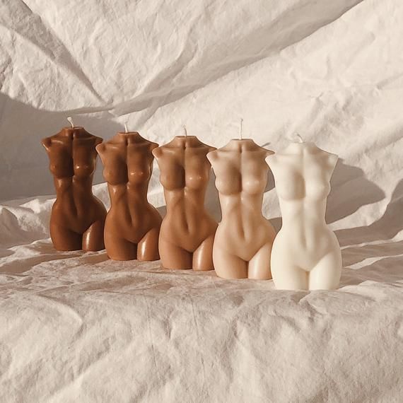 four candles in the shape of nude women on a white sheeted surface, with one candle lit