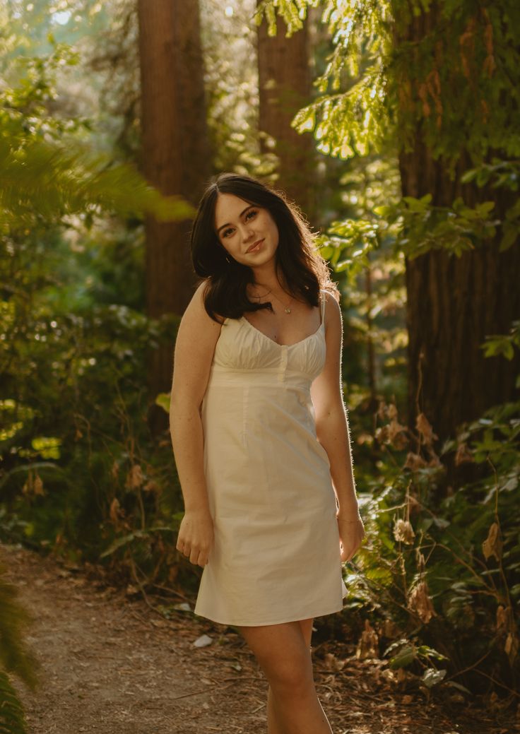 a woman in a white dress standing on a path through the woods with her legs crossed