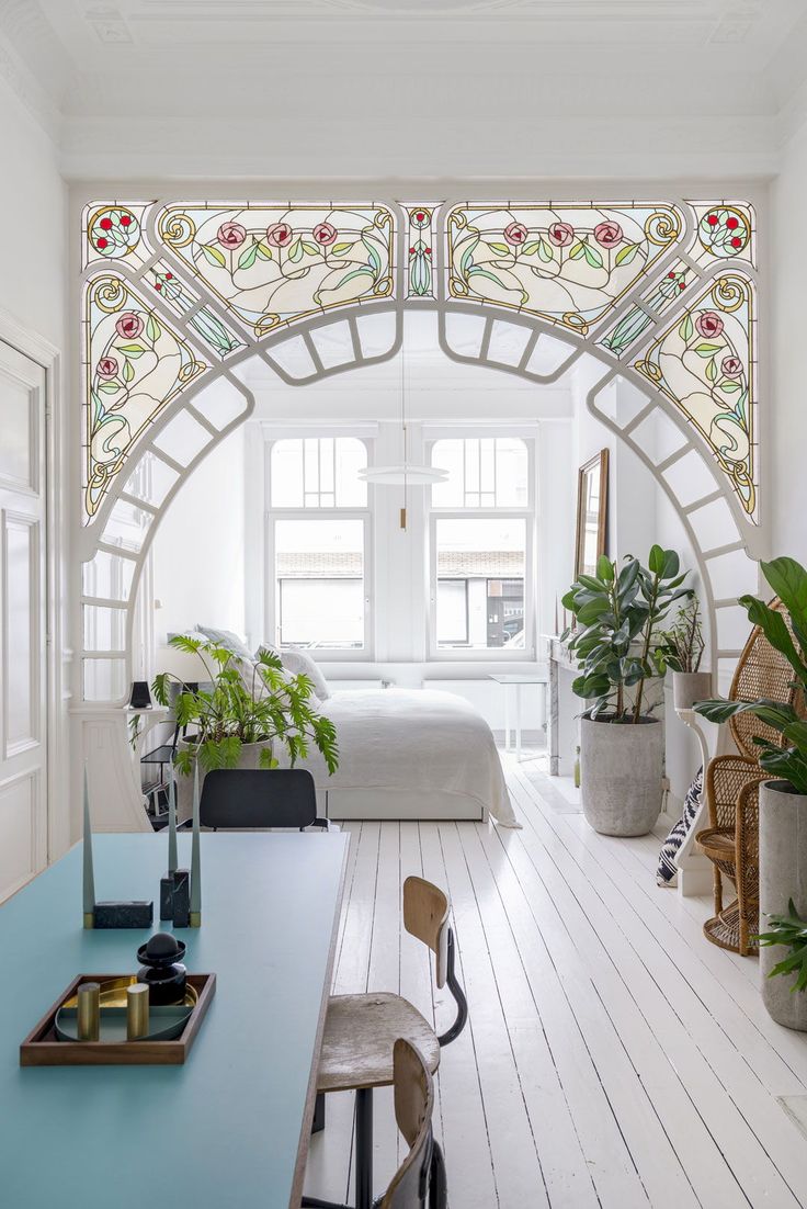 a bedroom with white walls and stained glass window above the bed, along with potted plants