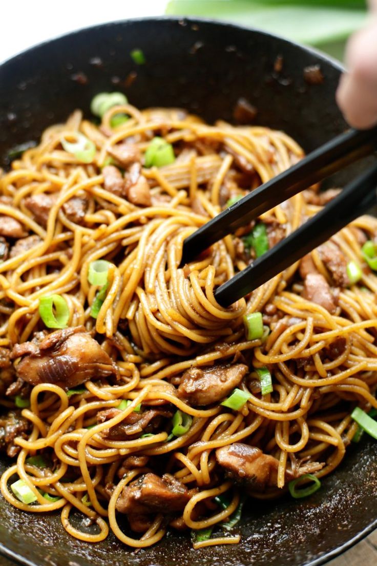 someone holding chopsticks over noodles in a skillet with meat and green onions