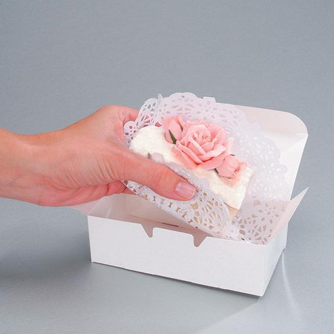 a hand is holding a piece of cake in a white box with lace and pink flowers