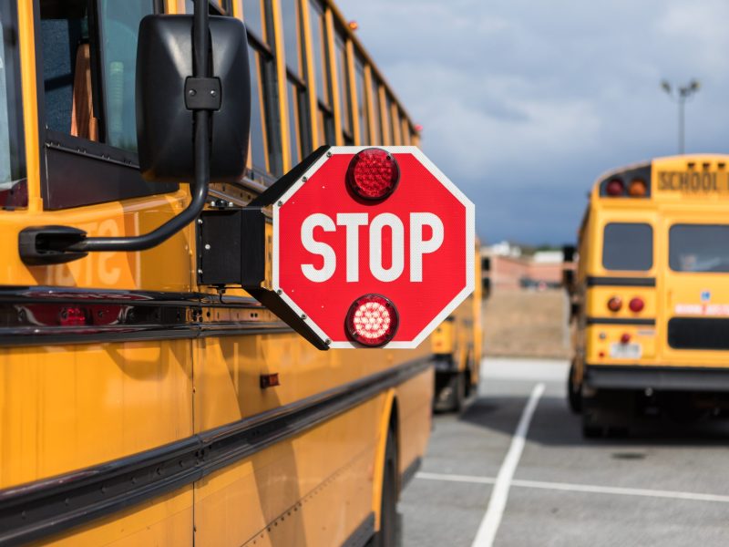 A yellow school bus in the foreground has its red STOP sign extended. In the background, two school buses are parked.