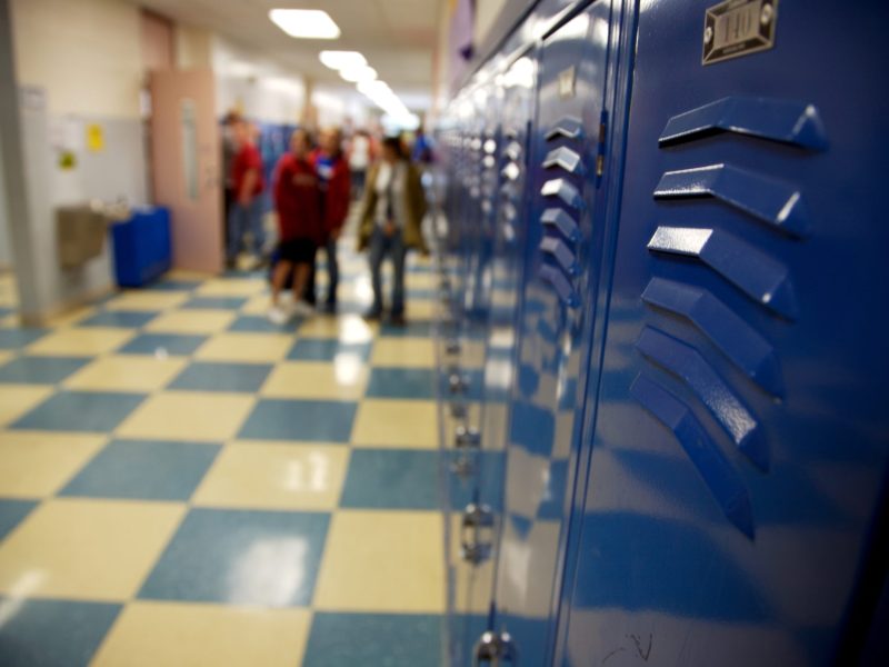 Blue school lockers are in focus in the foreground of the image showing a school hallway. In the background, out of focus, several people stand near each other and one person walks into a classroom.