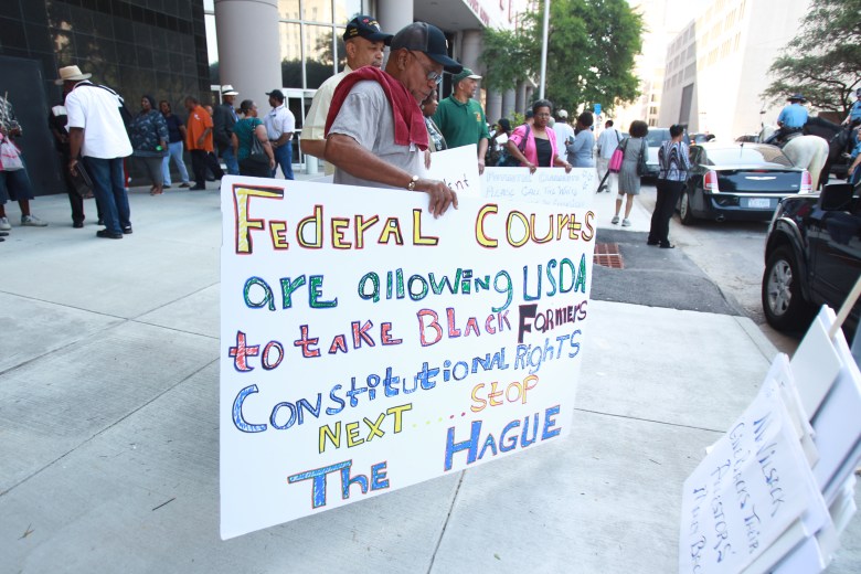 A man wearing a hat and glasses holds a protest sign that reads "Federal courts are allowing USDA to take Black Farmers constitutional rights. Next ... stop The Hague."