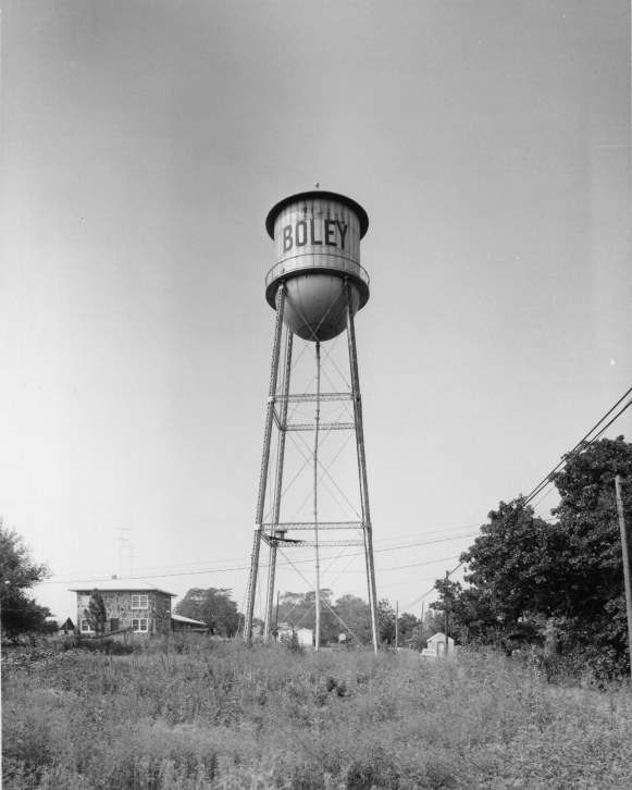 A black and white image of a water tower that has the word Boley on it. The water tower sits in an empty field. Behind it is a small building in the distanc.