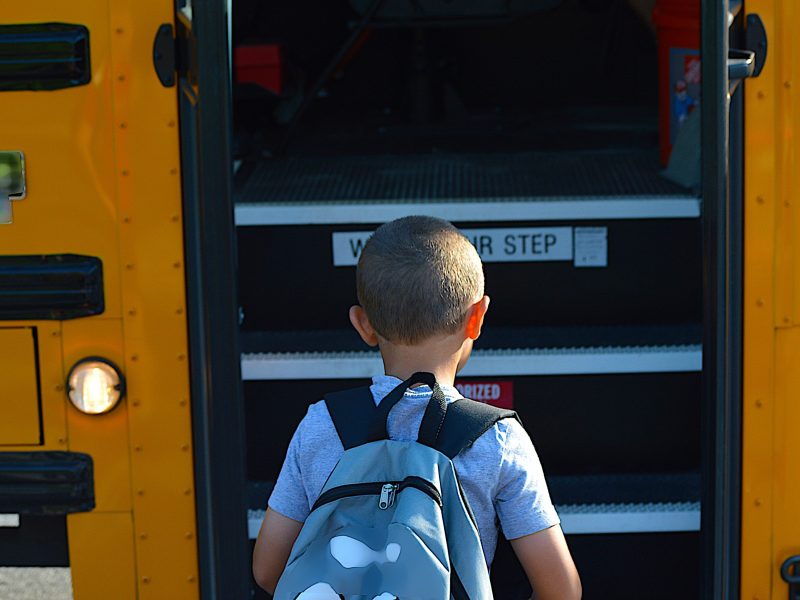 A child stands with their back to the viewer, in front of them are the steps leading into a school bus.