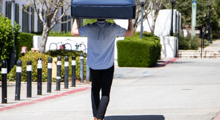 A student carrying a mattress on his head walks up a street.