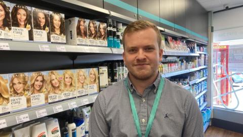 Harry Pearson is standing in front of a shelf full of hair dye in the pharmacy. He has short blond/brown hair and is wearing a grey shirt and a lanyard.