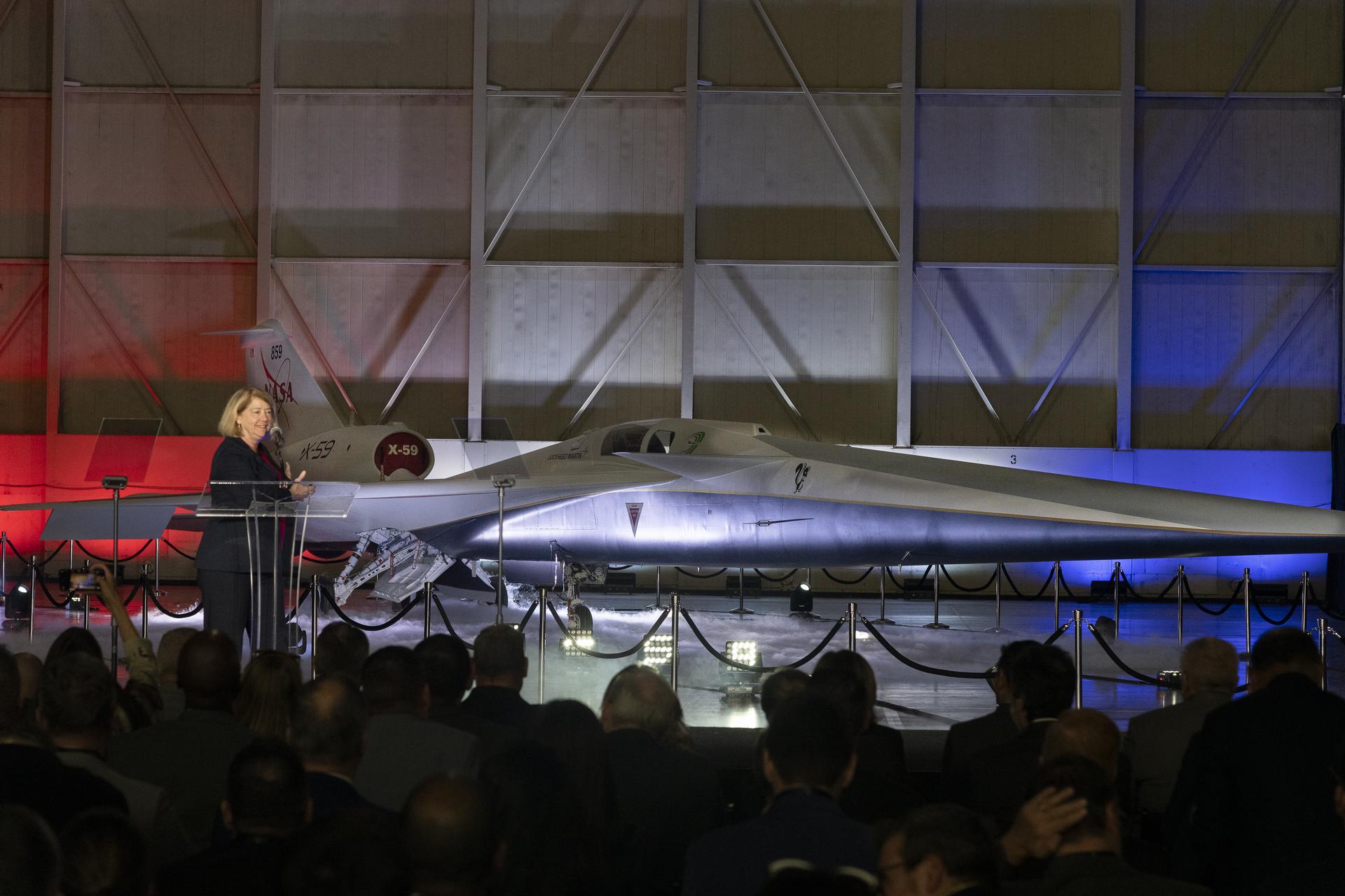 View of a lit stage inside a hangar. A woman speaks at a podium. In the immediate background, the X-59 sits, newly painted in its red, white, blue and gold livery.