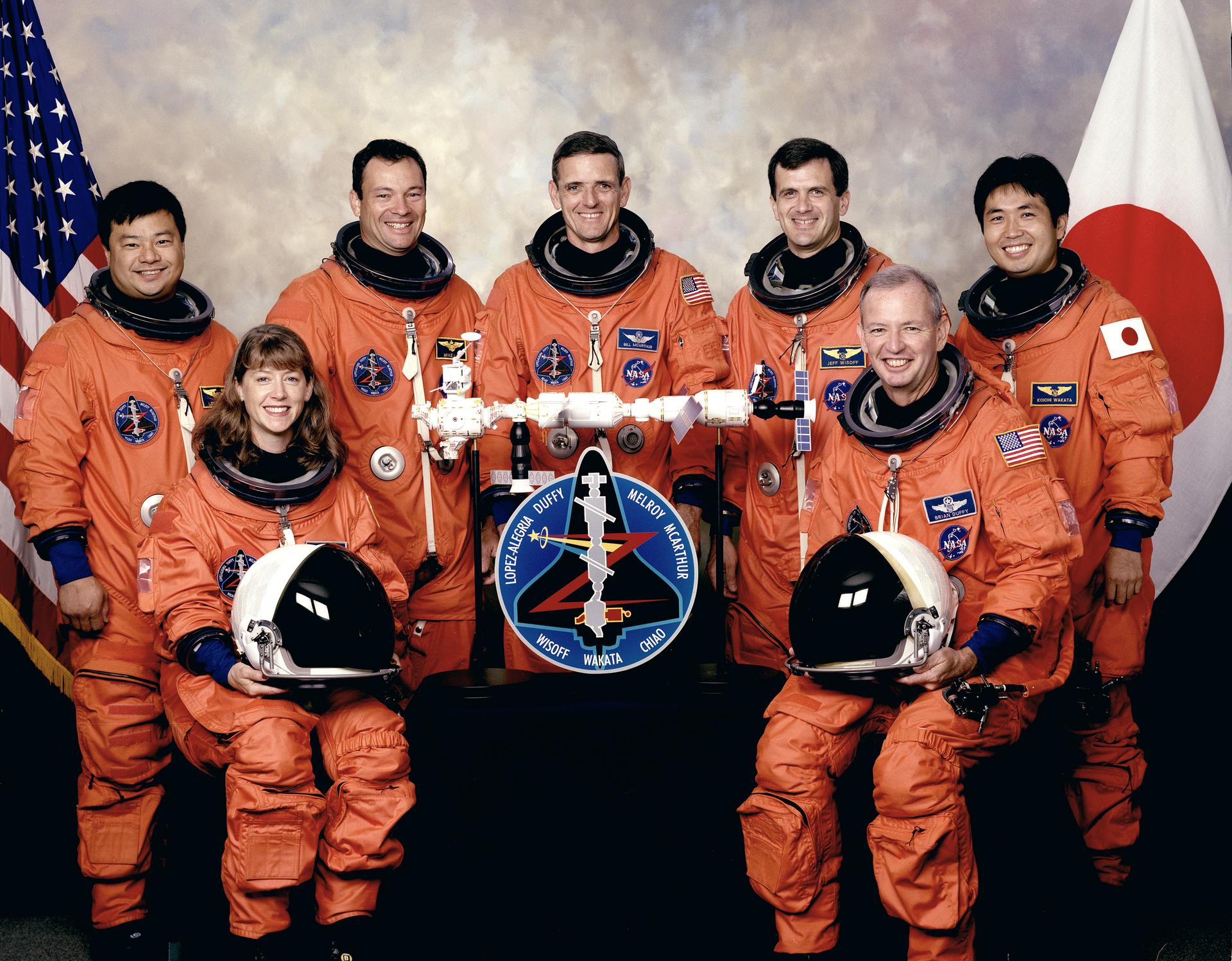 Seven astronauts in orange spacesuits pose in front of US and Japanese flag