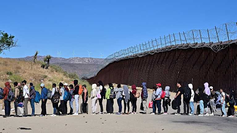 Ihmisiä seisoo jonossa Meksikon ja Yhdysvaltain rajalla. Raja-aita kiemurtaa kauaisuuteen.