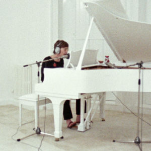 John Lennon soittaa valkoista flyygeliä, Yoko Ono istuu lattialla. Kuva dokumenttielokuvasta John & Yoko: Above Us Only Sky.