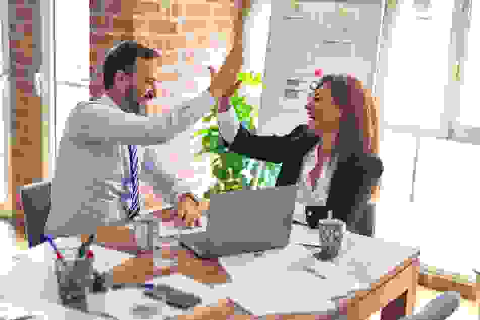 A man and woman dressed in corporate attire in an office sitting at a desk, giving each other a high five and smiling. Photo by krakenimages.
  