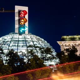 Eine Ampel vor dem Reichstagsgebäude leuchtet in einer Langzeitbelichtung in allen drei Phasen, wobei sich der Straßenverkehr als Leuchtspuren abzeichnet.