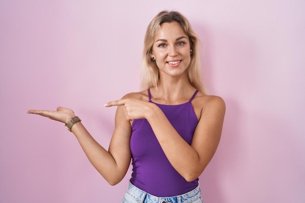 Free photo young blonde woman standing over pink background amazed and smiling to the camera while presenting with hand and pointing with finger.