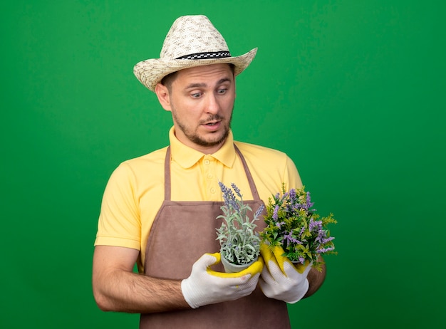 Бесплатное фото Молодой садовник в комбинезоне и шляпе в рабочих перчатках держит горшечные растения, глядя на них, смущенно стоя у зеленой стены
