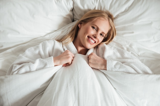 Young happy woman lying on big white bed with blanket