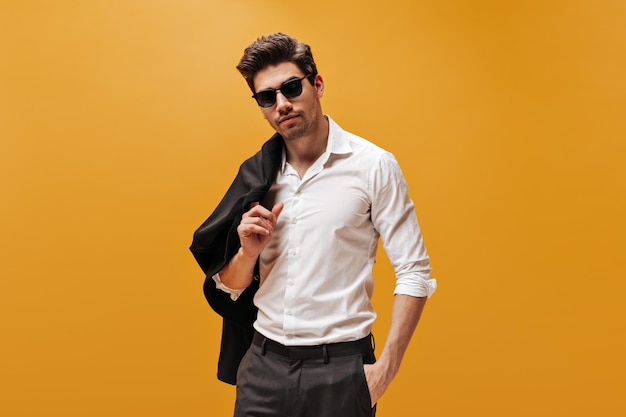 Привлекательный брюнет в стильной белой рубашке и модных солнцезащитных очках позирует на оранжевом фоне и держит черную куртку