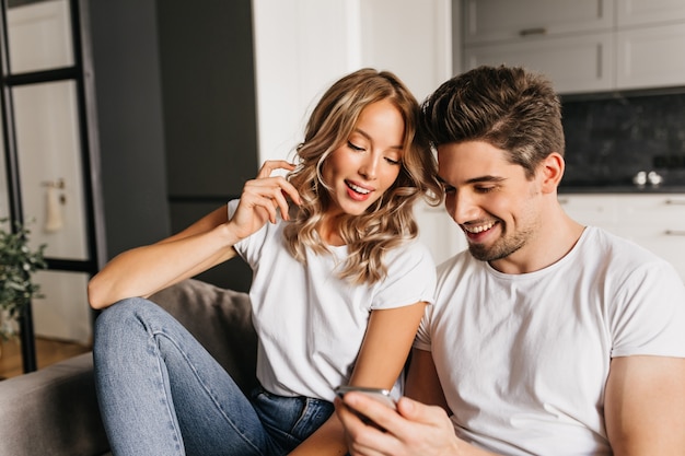 Счастливая пара с умным телефоном смотрит на экран и улыбается. Домашний портрет двух молодых людей, наслаждающихся днем вместе и читающих хорошие новости.