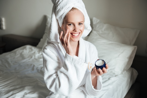 Happy smiling woman in bathrobe putting cream on face