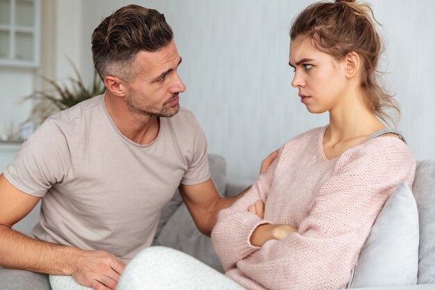 Side view of attentive man sitting on couch and calm down his serious girlfriend with crossed arms at home
