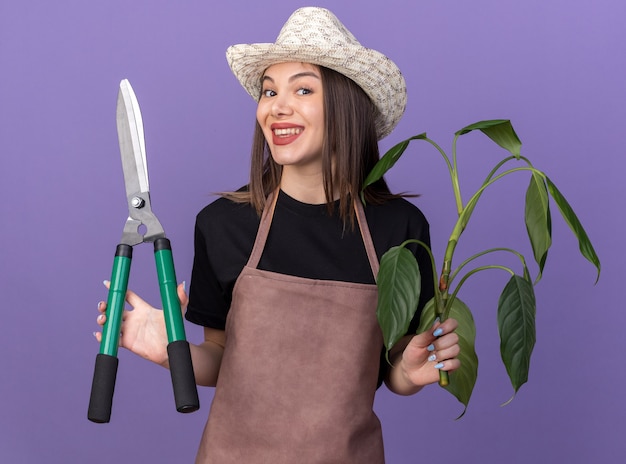 Бесплатное фото Улыбающаяся красивая кавказская женщина-садовник в садовой шляпе держит садовые ножницы и ветку растения, изолированную на фиолетовой стене с копией пространства