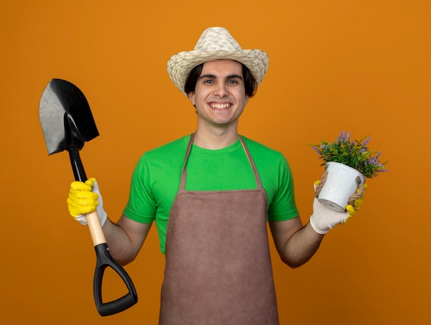 Улыбающийся молодой мужчина-садовник в униформе в садовой шляпе с перчатками держит лопату с цветком в цветочном горшке, изолированном на оранжевом