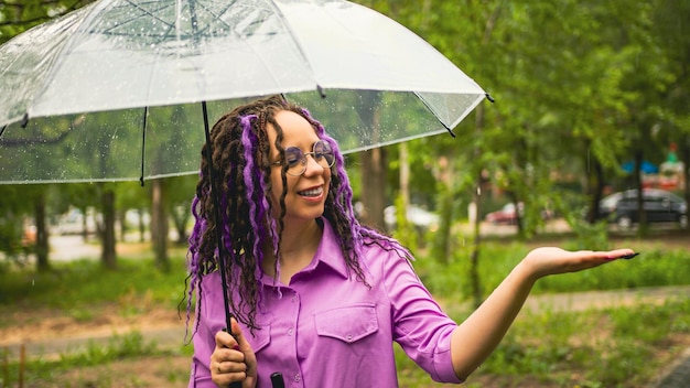 A woman with umbrella in hand in the rain Girl with glasses under an umbrella against the backdrop of an urban landscape Female with dreadlocks stands in a park with an umbrella