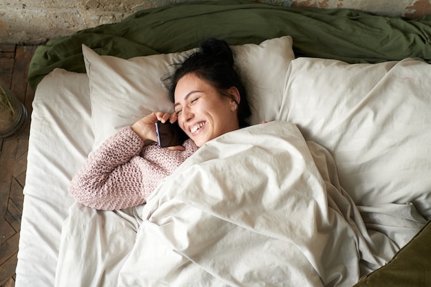 Young beautiful girl talking on a smartphone and laughs while lying in bed at home