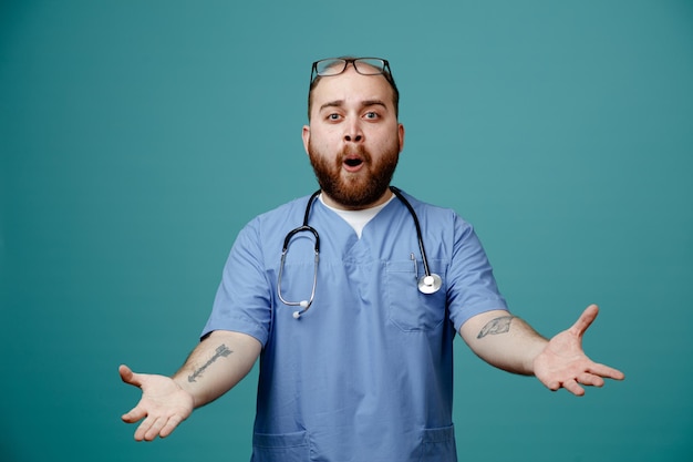 Бородатый мужчина-врач в форме со стетоскопом на шее в очках смотрит в камеру, счастливый и удивленный, раскинув руки в стороны, стоя на синем фоне