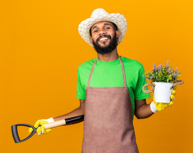 Уверенный молодой афро-американский парень садовник в садовой шляпе и перчатках держит лопату с цветком в цветочном горшке