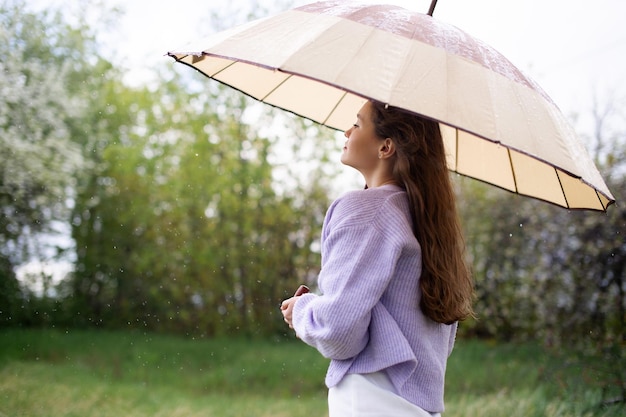 A girl in a sweater is standing under an umbrella it's raining
