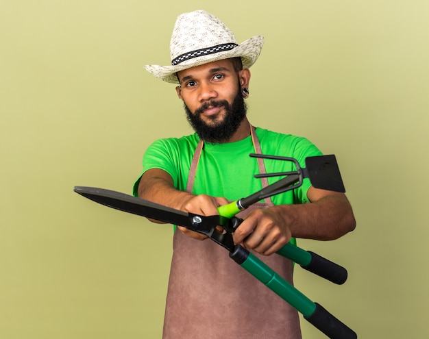 Довольный молодой афро-американский парень садовник в садовой шляпе протягивает ножницы с граблями для мотыги перед камерой