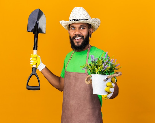 Улыбающийся молодой афро-американский парень садовник в садовой шляпе и перчатках держит лопату с цветком в цветочном горшке, изолированном на оранжевой стене