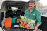 A smiling man stands in front of a van. He is holding a box of friot and vegetables.