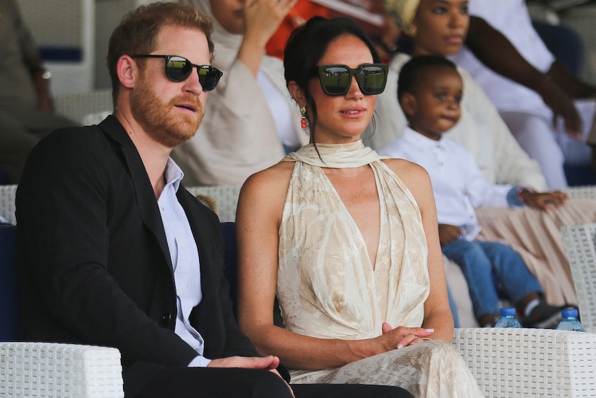 Prince Harry wearing a suit and sunglasses leans in to Meghan Markle wearing sunglasses and a yellow dress.