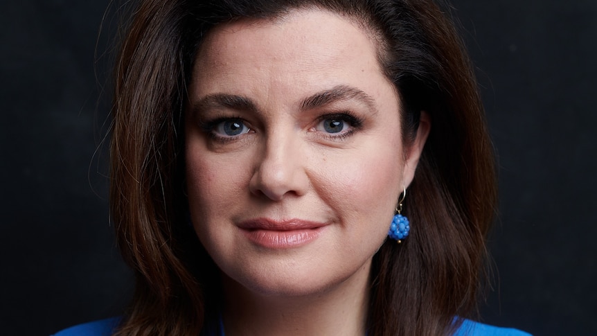 A white woman with long dark brown hair wearing a blue suit and matching blue earrings