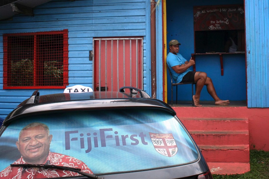 An election poster for "Frank" Bainimarama can be seen in the rear window of a taxi as a man gestures from a doorway to gym.