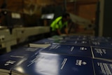 A close up of 2024-25 federal budet papers stacked on a crate, with a printmaker in the background